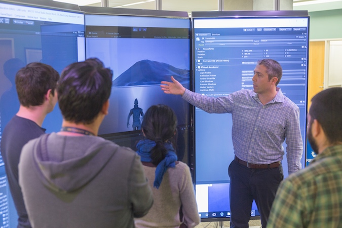 A man points to a three large monitors while four students face him and listen to his explanation.