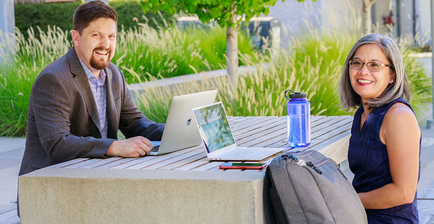 Joel Spencer and Jennifer Manilay smile from an outdoor table on campus